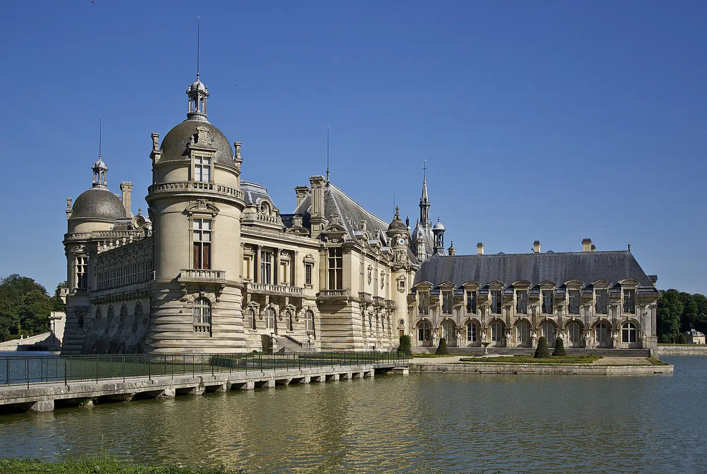 Le château de Chantilly (Oise) au bord de son plan d'eau, avec sa tour à dôme et son aile à arcades, sous un ciel bleu