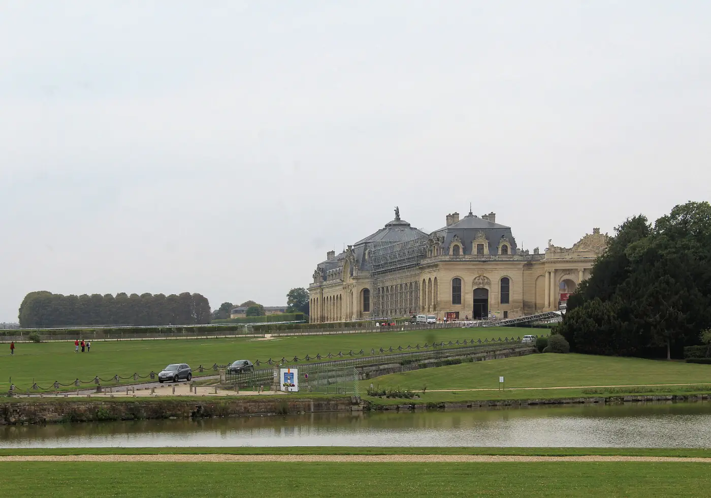 Les Grandes Écuries du domaine de Chantilly vues depuis le parc, avec une pièce d'eau et une allée où circulent des voitures