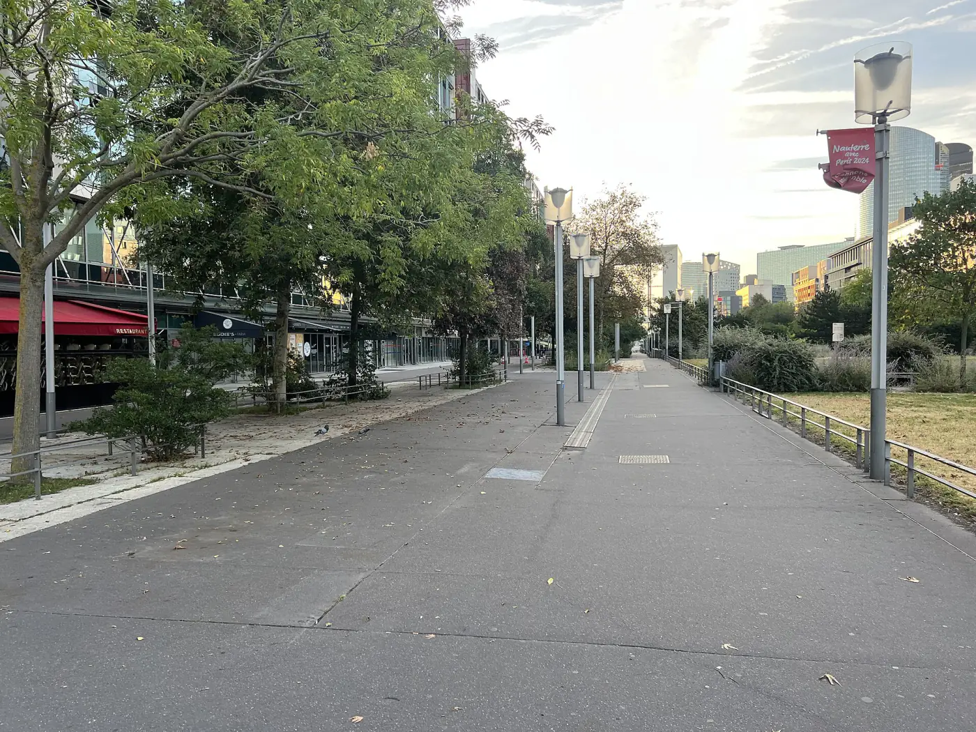Terrasse de l'Arche in Nanterre: a pedestrian walkway lined with trees and lamp posts, with the La Défense towers in the background.